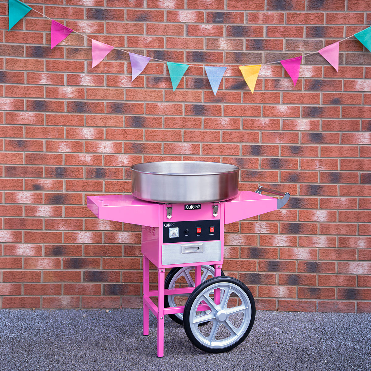 Máquina de algodão doce rosa com detalhes prateados, posicionada em frente a uma parede de tijolos, decorada com bandeirinhas coloridas