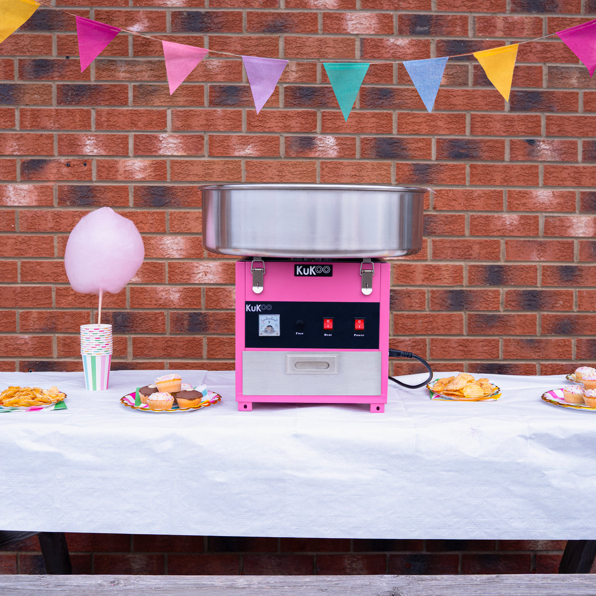 Máquina de algodão doce rosa com acabamento em aço inox, decorada com bandeirinhas coloridas, acompanhada de doces e cupcakes em uma mesa branca