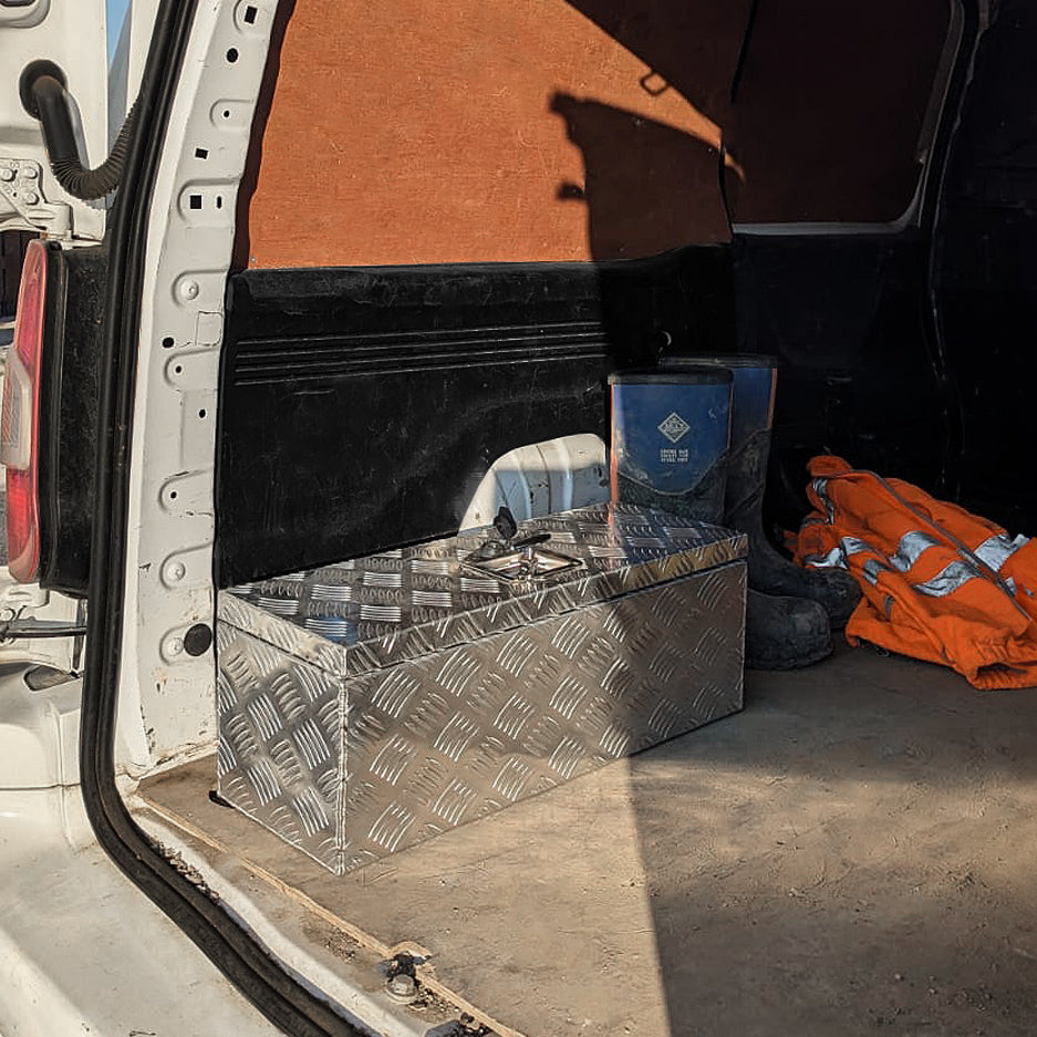Caixa de ferramentas de alumínio 35L com textura em relevo, prateada, posicionada no porta-malas de uma van, ao lado de ferramentas e roupas de trabalho
