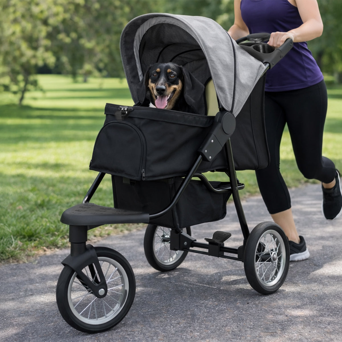 Carrinho para animais de estimação cinza com estrutura preta, rodas grandes, e um cão feliz dentro, em um parque verde