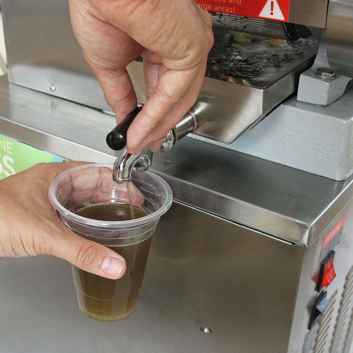 Mão segurando um copo transparente enquanto suco de cana fresco é dispensado de uma máquina de aço inoxidável brilhante