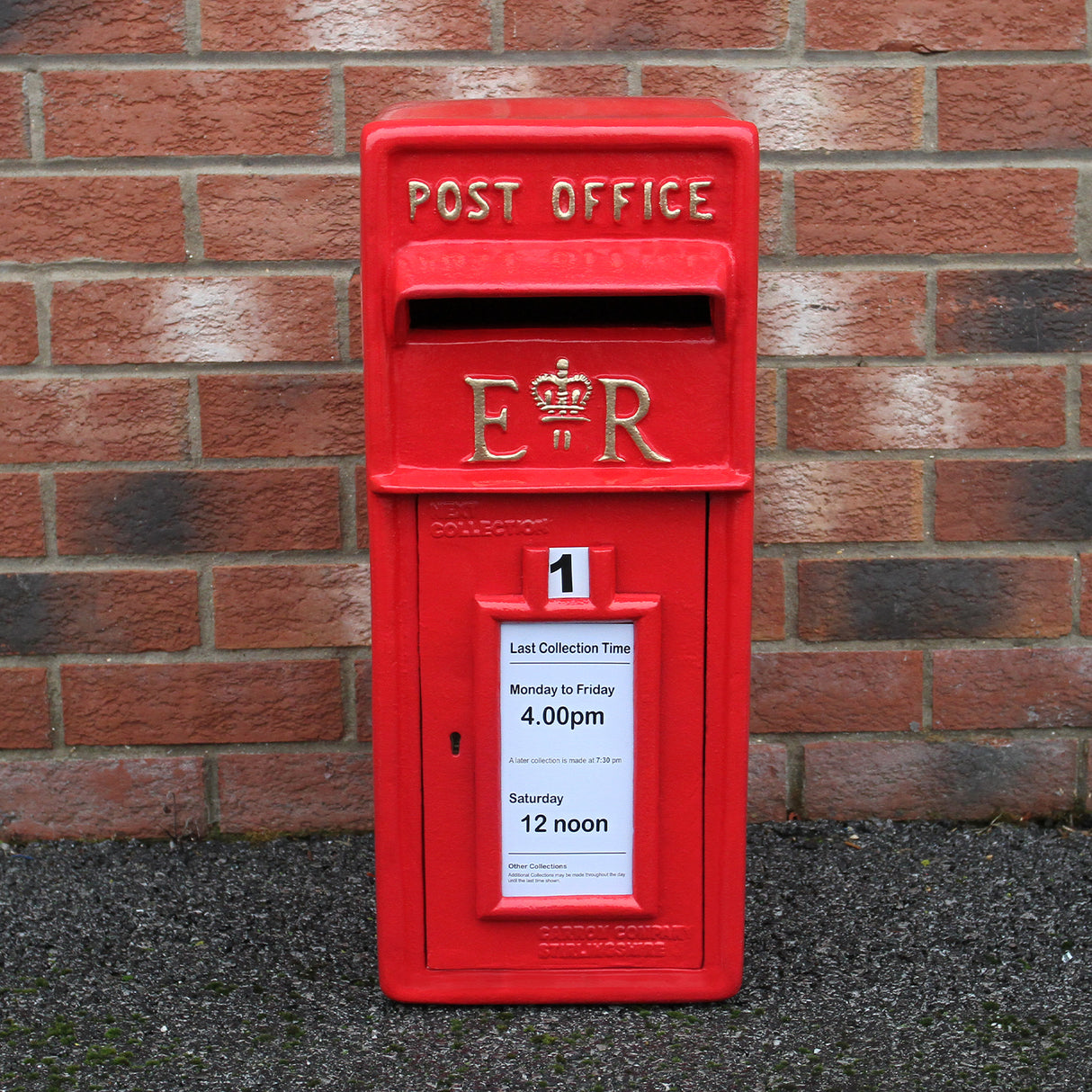 Caixa de correio vermelha Royal Mail com suporte, destacando detalhes em relevo e aviso de horários de coleta em papel branco