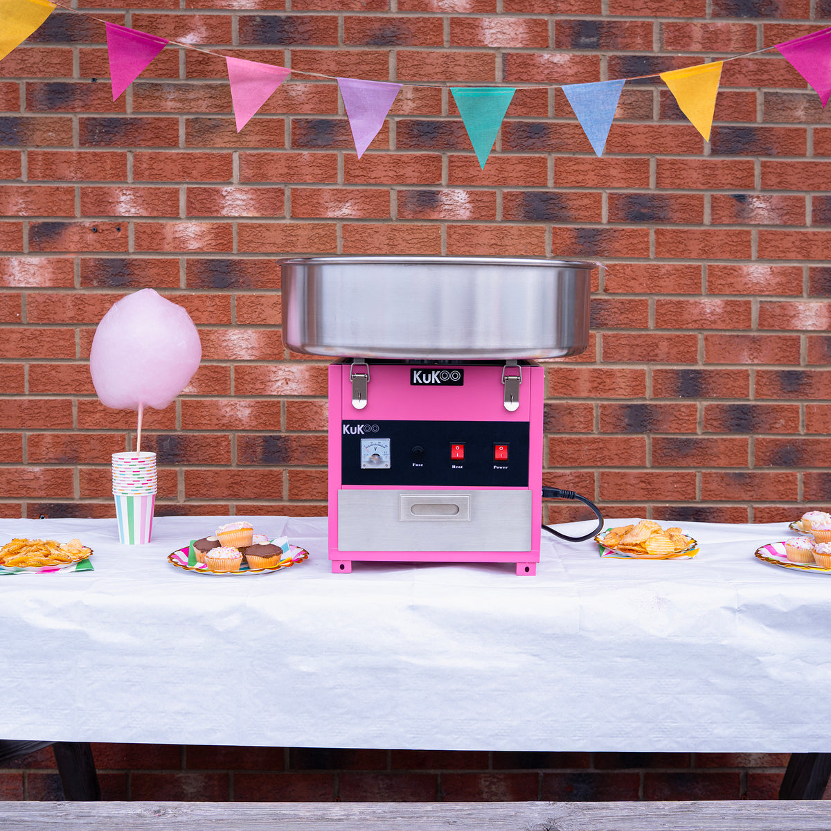 Máquina de fazer algodão doce KuKoo em rosa e prata, com tigela metálica, decorada com bandeirinhas coloridas e doces ao fundo