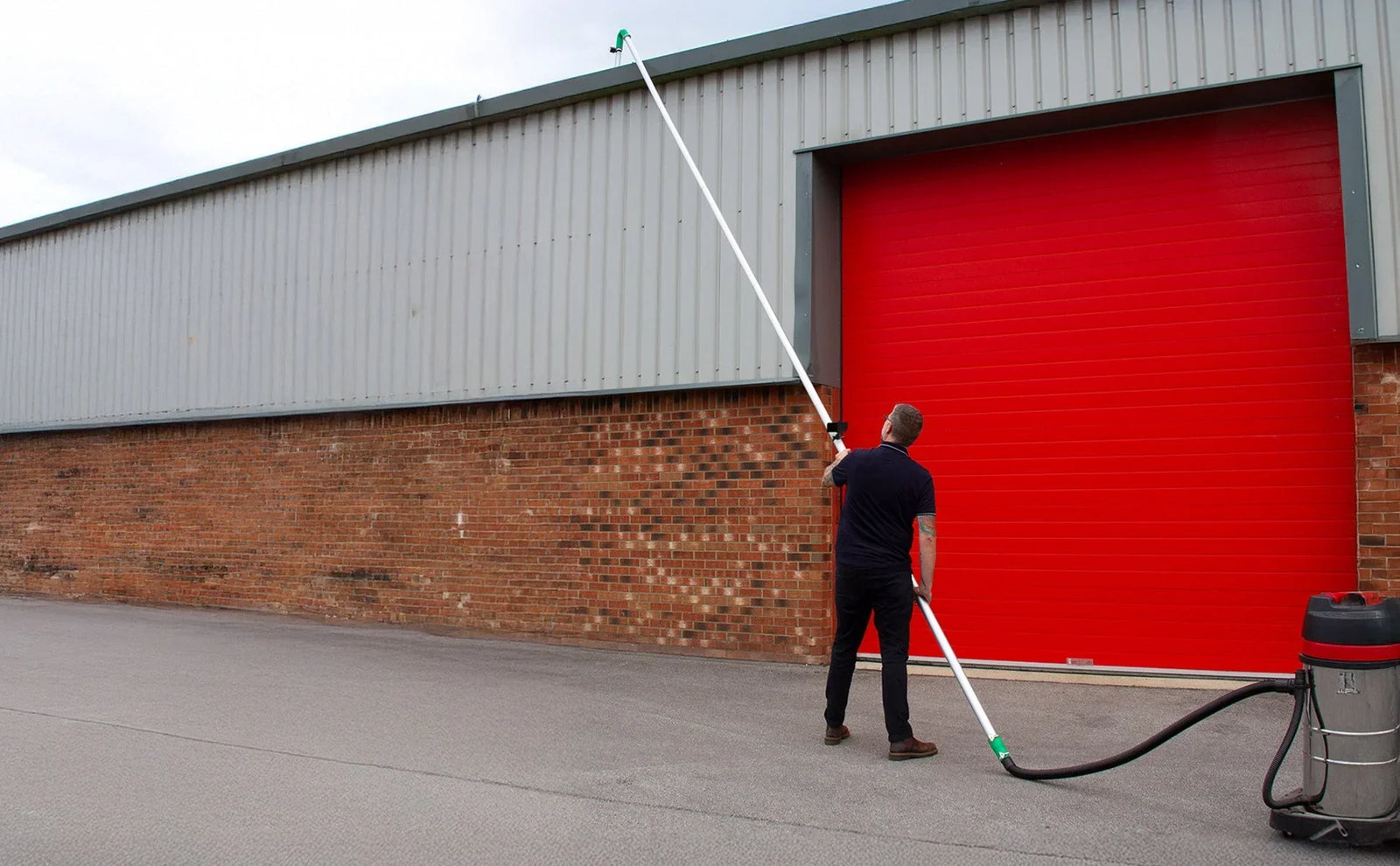 Homem usando um aspirador industrial com tubo longo para limpar calhas, em frente a um galpão com porta vermelha e parede de tijolos