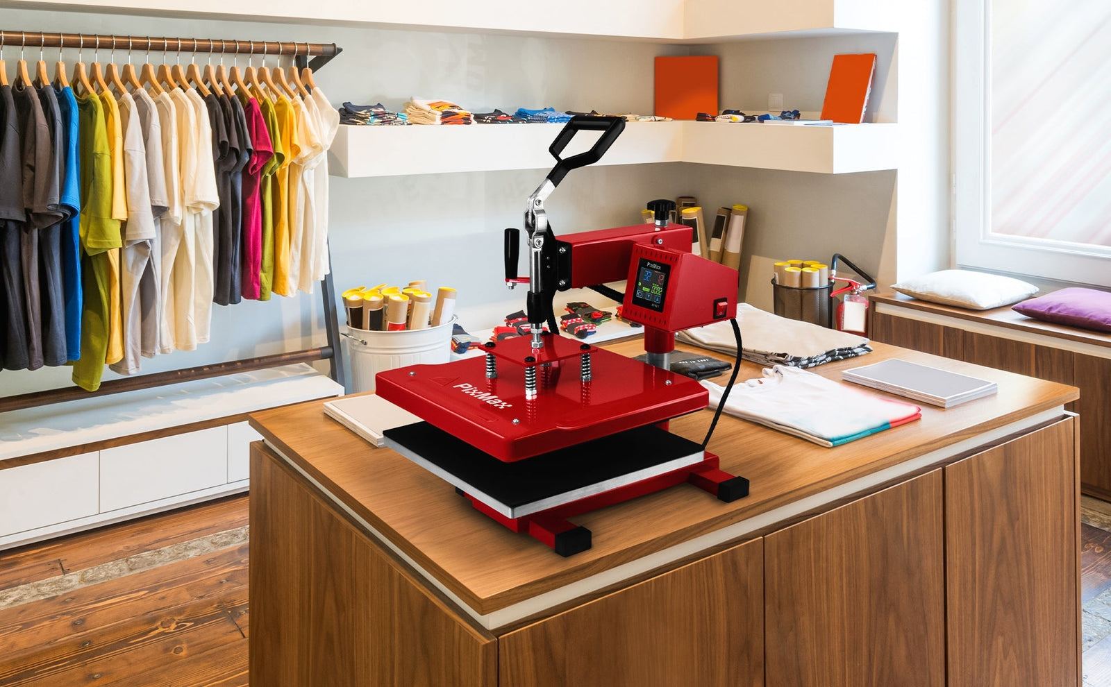 Prensa térmica vermelha sobre mesa de madeira, com camisetas coloridas penduradas ao fundo e materiais de artesanato organizados