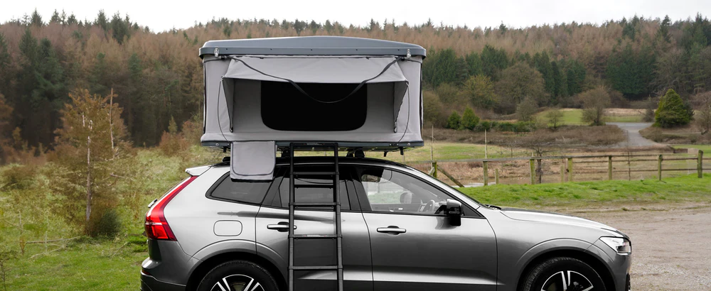 Tenda de teto cinza montada em um carro prateado, com vista para uma paisagem verde e montanhosa ao fundo, ideal para acampamentos