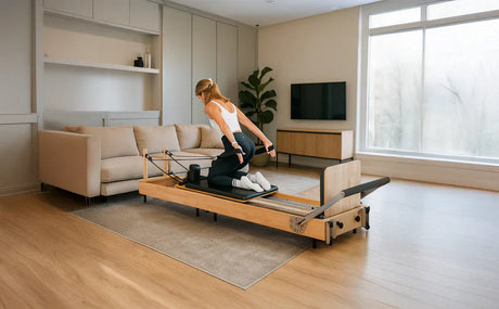 Mulher praticando Pilates em um reformer de madeira clara, em sala moderna com sofá e janela grande iluminando o ambiente