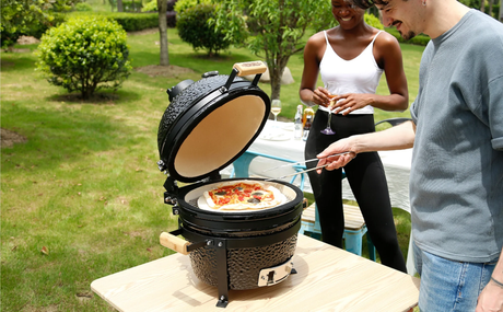 Churrasqueira Kamado preta com tampa aberta, pizza colorida sendo assada, pessoas sorrindo em um ambiente ao ar livre com verde ao fundo