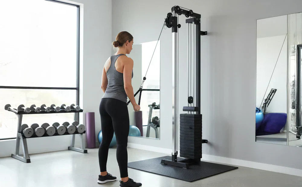 Mulher em leggings pretas e top cinza usando uma máquina de cabos de parede em um ambiente de academia moderno e iluminado