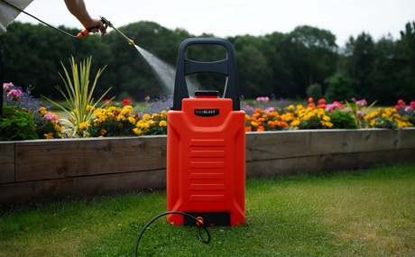 Pulverizador de jardim laranja vibrante em primeiro plano, com flores coloridas ao fundo e um jardineiro borrifando água