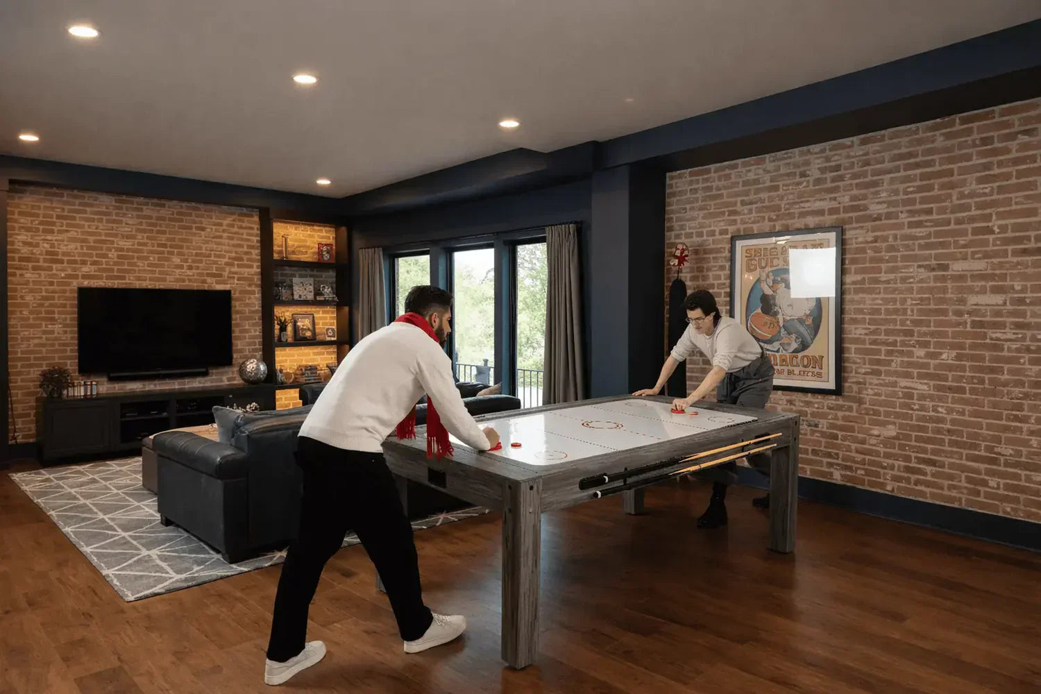 Dois homens jogando em uma mesa de jogos 7 em 1, com estrutura metálica e superfície branca, em uma sala moderna com paredes de tijolos expostos