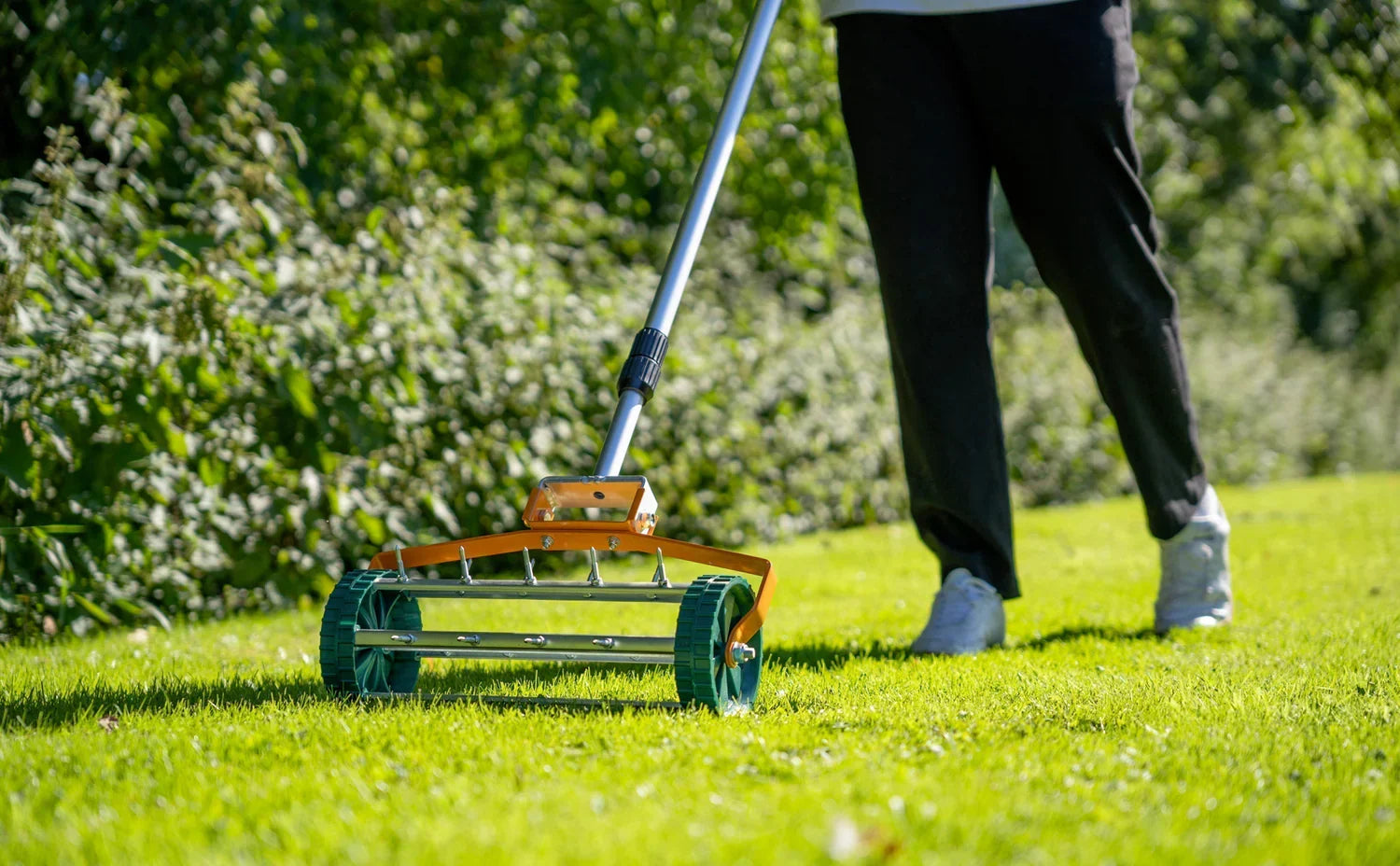 Um cortador de grama manual laranja e verde sendo usado em um gramado verde vibrante, com uma pessoa caminhando ao fundo