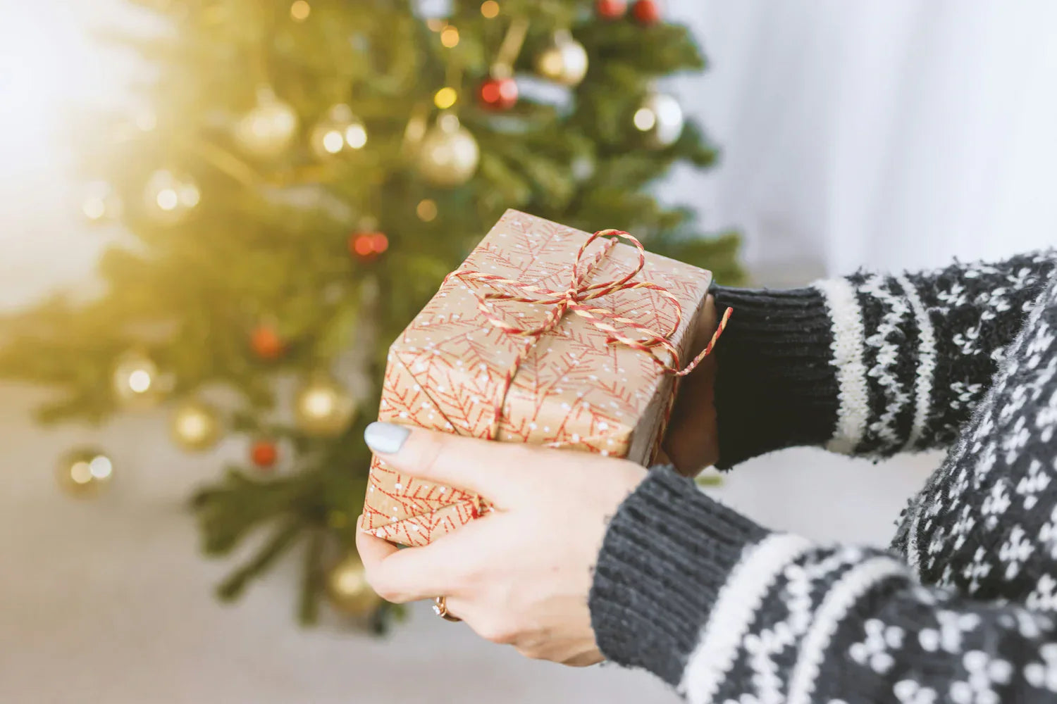 Caixa de presente decorada com papel dourado e laço vermelho, segurada por mãos, com árvore de Natal ao fundo iluminada