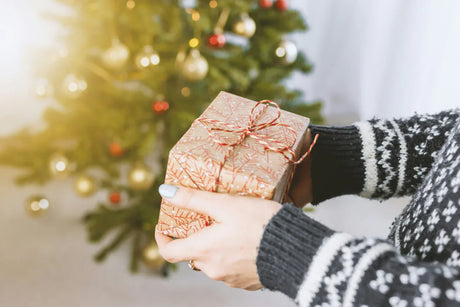Caixa de presente decorada com papel dourado e laço vermelho, segurada por mãos, com árvore de Natal ao fundo iluminada