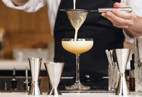 Coquetel cremoso sendo coado em taça elegante, com utensílios de bar prateados e fundo de madeira clara, criando um ambiente sofisticado