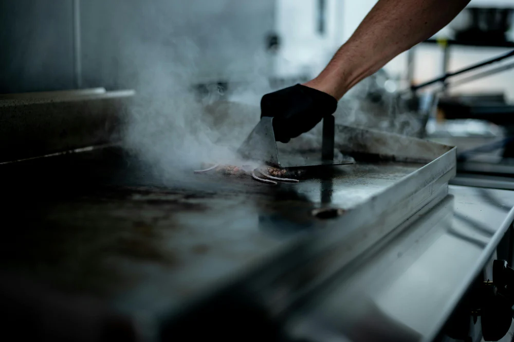 Chapa elétrica com carne sendo grelhada, vapor subindo, mão usando espátula, fundo de cozinha profissional com utensílios