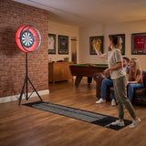Jogo de dardos com moldura LED em destaque, com luzes vermelhas, em ambiente aconchegante com sofá e mesa de bilhar ao fundo
