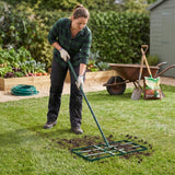 Nivelador de relva verde de 100 cm sendo usado por uma mulher em um gramado, com textura de solo solto ao redor e ferramentas de jardinagem ao fundo
