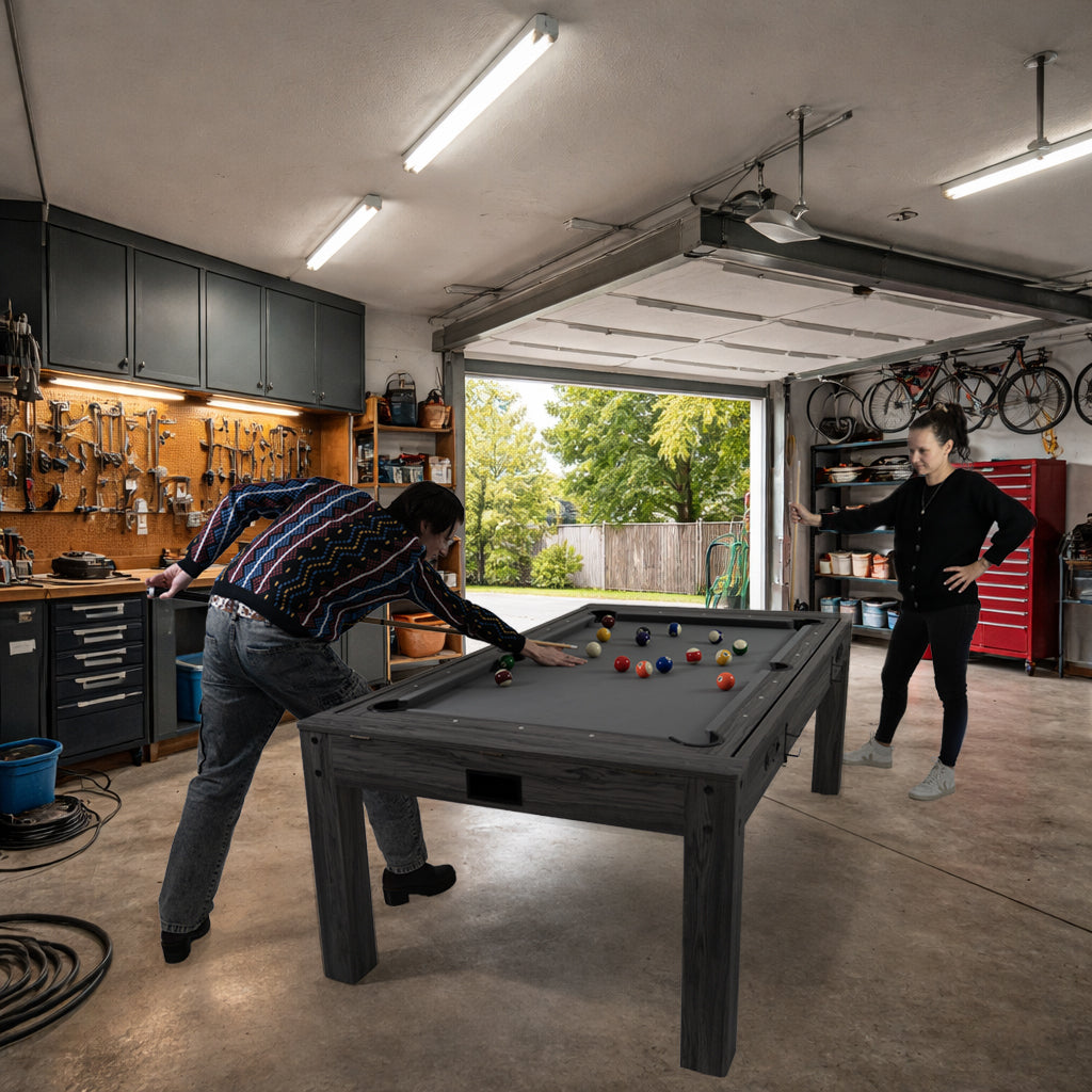Mesa de jogos 7 em 1 cinza, com superfície de feltro, dardos e luz LED, em ambiente de garagem com ferramentas e bicicletas ao fundo