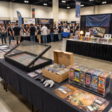 Mesa de convenção com cartas colecionáveis organizadas em caixas, controle de videogame e público ao fundo em ambiente iluminado