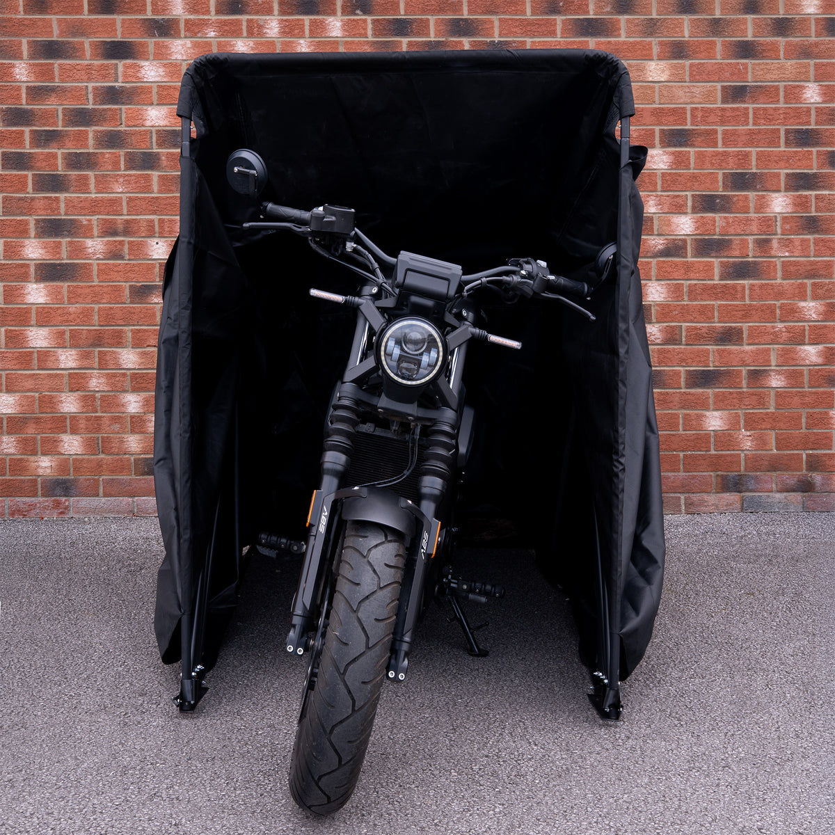 Motocicleta preta com detalhes em metal, parcialmente coberta por uma capa preta, em frente a uma parede de tijolos vermelhos