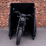 Motocicleta preta com detalhes em metal, parcialmente coberta por uma capa preta, em frente a uma parede de tijolos vermelhos