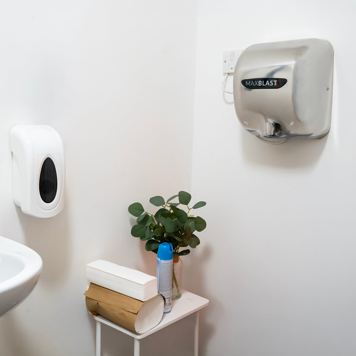 Secador de mãos prateado montado na parede, dispenser de sabonete branco ao lado, planta verde em vaso sobre mesa branca, spray azul próximo