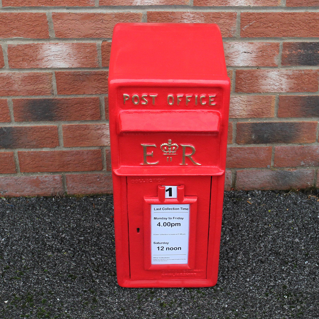 Caixa de correio Royal Mail vermelha com suporte, destacando detalhes em dourado e placa de horários de coleta em papel branco