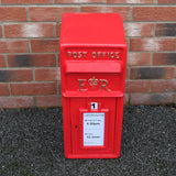 Caixa de correio Royal Mail vermelha com suporte, destacando detalhes em dourado e placa de horários de coleta em papel branco