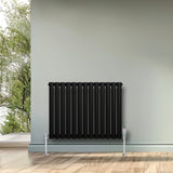 Radiador de coluna simples oval preto, com válvulas, instalado em parede cinza, destacando seu design moderno e elegante