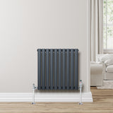Radiador de coluna simples oval cinza antracite, com tubos verticais e suportes metálicos, instalado em parede clara, próximo a sofá claro