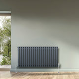Radiador de coluna simples oval cinza antracite, com 600 mm de altura e 1200 mm de largura, instalado em parede clara com piso de madeira