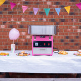 Máquina de fazer algodão doce KuKoo em rosa e prata, com tigela metálica, decorada com bandeirinhas coloridas e doces ao fundo