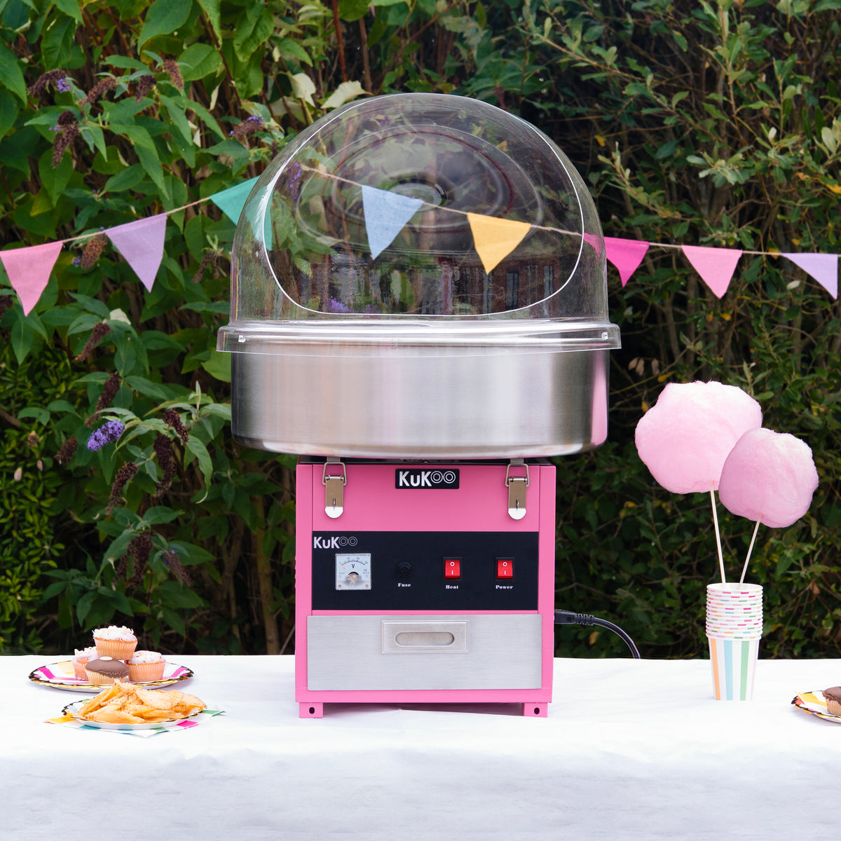 Máquina de algodão-doce KuKoo em rosa com cúpula protetora, acompanhada de algodão-doce e bandejas coloridas em um ambiente festivo