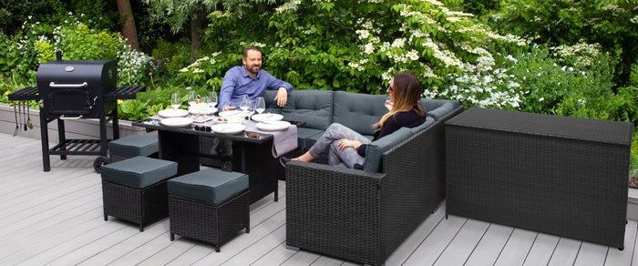 Conjunto de móveis de jardim em vime preto, com sofás confortáveis, mesa de vidro e churrasqueira, cercado por vegetação verde exuberante